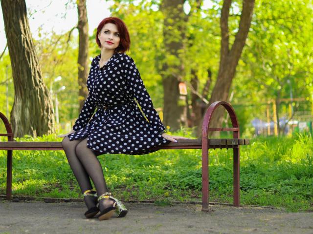 Lisa, con i capelli rossi, un vestito nero a pois, calze nere e scarpe nere, seduta su una panchina di legno in un parco, con le gambe incrociate, che guarda verso la fotocamera..