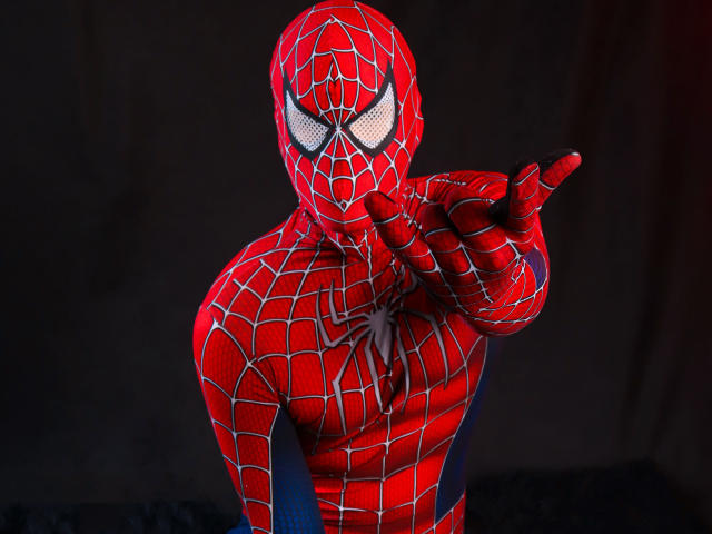 SebastiianMaxime in een rood Spider-Man kostuum met witte webpatronen, wijzend met de rechterhand, met een zwarte oogmasker, tegen een donkere achtergrond. Het rode kostuum bedekt het lichaam van SebastiianMaxime..