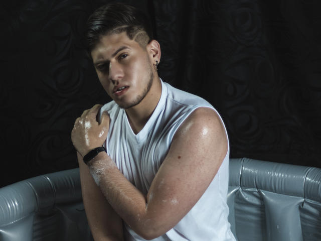 SebastiianMaxime mit glänzenden, verschwitzten Armen, der ein weißes, ärmelloses Hemd, ein schwarzes Armband und einen Ohrring trägt, blickt mit einem leichten, verführerischen Lächeln in die Kamera. Dunkler Hintergrund, ein grauer, runder Sitz..