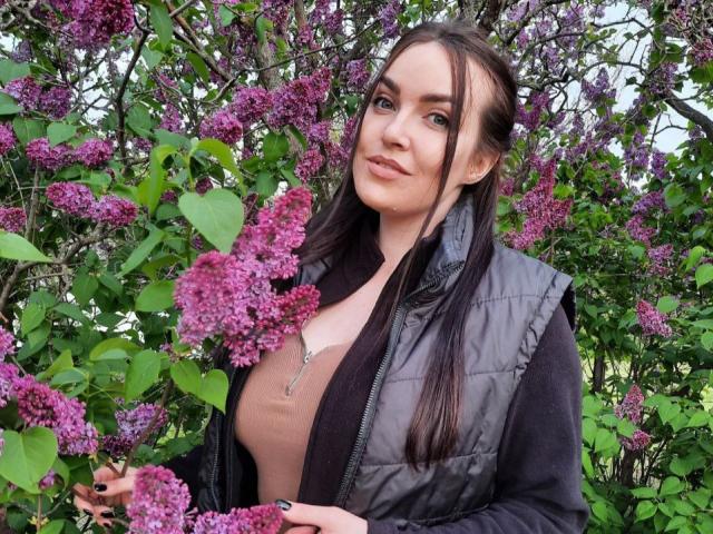 WilsonMelanie, con dei lillà viola, indossa un gilet imbottito nero, una maglietta rosa e una collana, ha lunghi capelli castani, pelle chiara e un sorriso delicato, ed è in piedi in un cespuglio verde e rigoglioso..