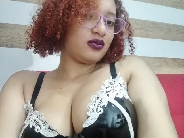 VioletaSwitch, con i capelli rossi ricci, rossetto viola, occhiali, reggiseno di pizzo nero e bianco, che mette in mostra un seno prosperoso, seduta su un divano rosso..