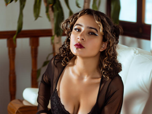 AlexiaBloom with curly brown hair, wet skin, dark lipstick, and black lace top, showing her tits, looking up with blue eyeliner, sitting on a white couch.