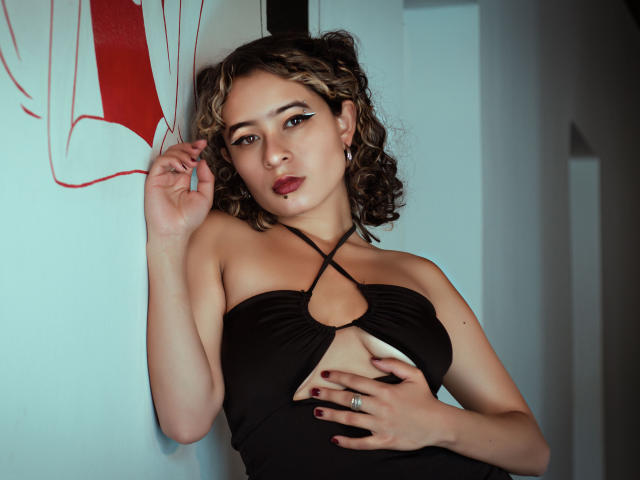 AlexiaBloom with curly brown hair, black cutout dress, and black eyeliner, holding her tits, wearing a ring, and looking at the camera, standing against a white wall.