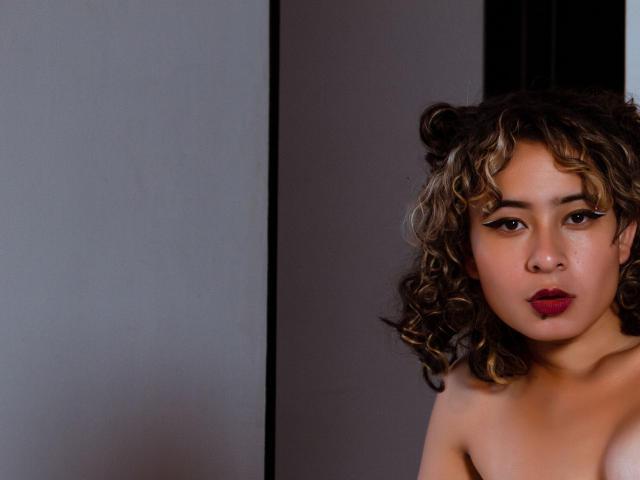 AlexiaBloom has curly brown hair, dark red lipstick, and thick eyeliner, staring with a neutral expression, topless, right side of the image, against a dark and light background.