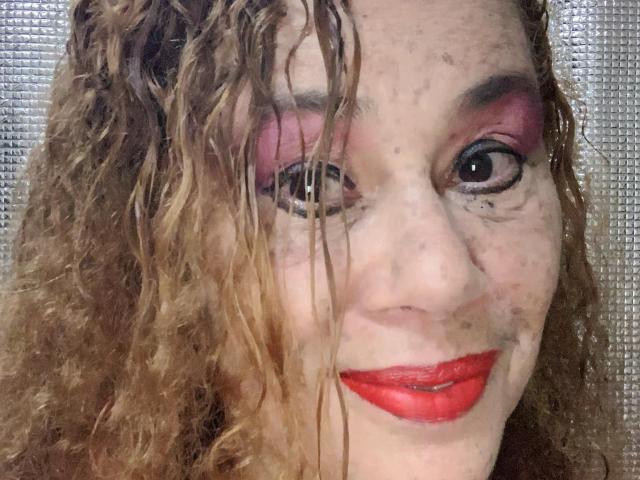 MadelaineLu tiene el cabello castaño rizado, lápiz labial rojo, sombra de ojos rosa, y un rostro ligeramente despeinado con pecas visibles y maquillaje de ojos oscuro, sonriendo a la cámara..