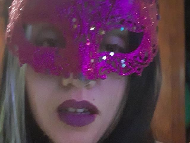HotNata indossa una maschera color viola con glitter, rossetto scuro e i capelli biondi sono parzialmente visibili, mostrando il naso e le labbra. I suoi occhi sono nascosti dalla maschera..