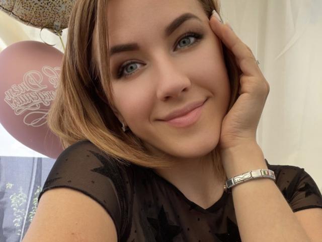 Misstyk with short blonde hair, blue eyes, and a cute smile, wearing a black star-patterned top, touching her cheek, and a silver watch on her wrist.