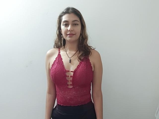 RosalieBlack, con i capelli castano ondulati, indossa una camicetta di pizzo rossa che mette in mostra il seno, una gonna nera e una collana, ed è in piedi di fronte a un muro bianco liscio, sorridendo leggermente..