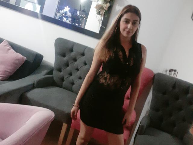RosalieBlack, in un abito nero di pizzo, che mette in mostra i suoi piccoli seni e glutei, è in piedi in una stanza con sedie grigie e rosa, sorridendo leggermente, con lunghi capelli castani..