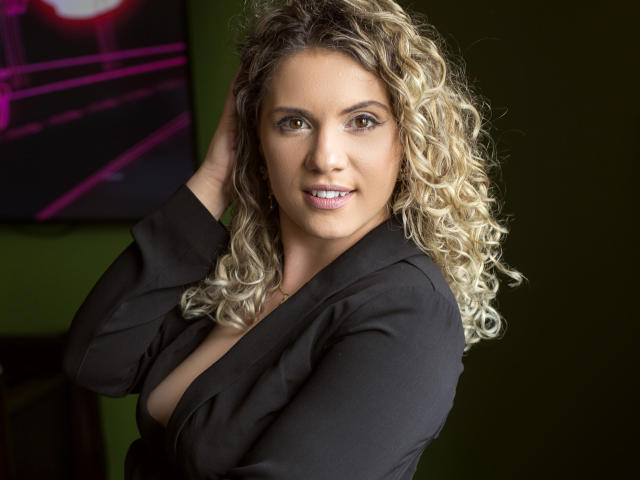 LucilleXSugar with curly blonde hair, wearing a black shirt, has her hand in her hair, showing her tits slightly, smiling, green background, pink neon art in the top left.