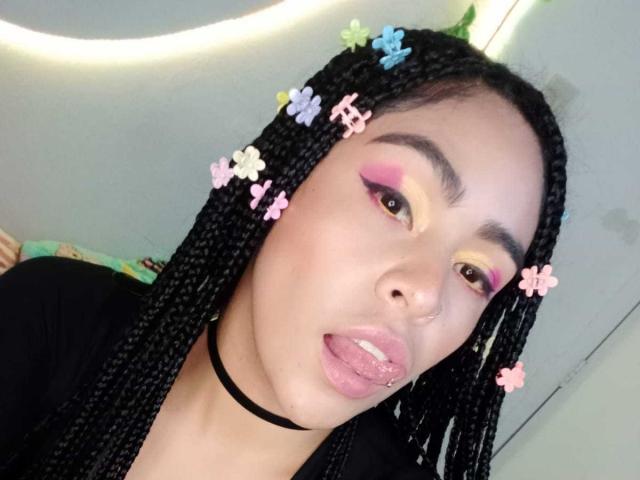 StellaBeckker has braided black hair with pink, blue, and white flower clips. She's sticking out her pink glittered tongue, wearing pink and yellow eye makeup, and a black choker.