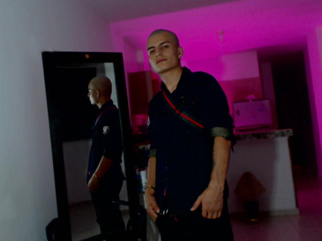 ShatSex, careca, vestindo uma camisa azul marinho escura com uma alça vermelha, está em frente a um espelho, mostrando seu reflexo. Uma luz rosa projeta um brilho suave..