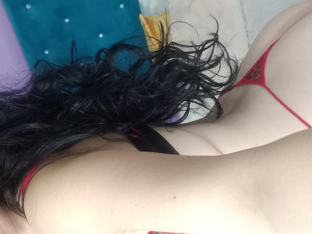 ValentinaAnal with black wavy hair, pale skin, red thong, and visible butt, lying on a bed, showing her back and butt. Her thong has a leopard print pattern.