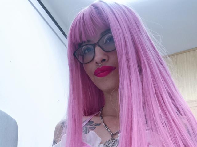 LailaStewart with pink hair, glasses, red lipstick, tattoos, and a necklace, showing off her tits in a selfie.