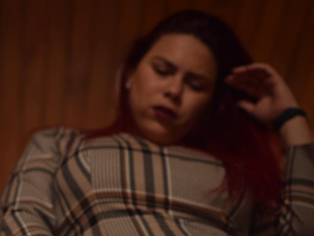 Salma Brown mit roten Haaren, verschwommenem Gesicht, trägt ein kariertes Hemd, hat eine Hand im Haar, blickt nach unten, weiches Licht, brauner Holz Hintergrund. Ihre Brüste sind vom Hemd bedeckt..