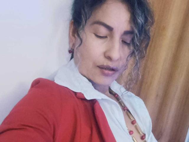 EroticSasha met krullend donker haar, die een rode cardigan draagt over een witte blouse, heeft een ketting met rode kralen, haar ogen zijn gesloten, ze heeft een lichte glimlach, op de achtergrond is hout te zien..