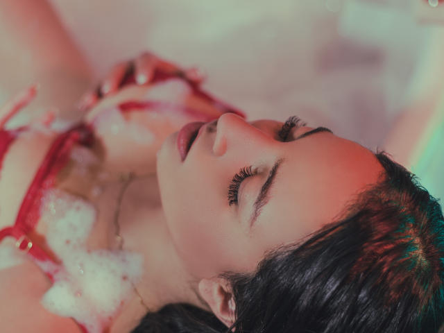 GiannaRusell, les yeux fermés, portant de la lingerie rouge, avec des bulles sur la poitrine, des cheveux foncés et mouillés, un éclairage rose et vert, un maquillage léger, une expression détendue, un petit anneau au nez, et un rouge à lèvres légèrement entrouvert..