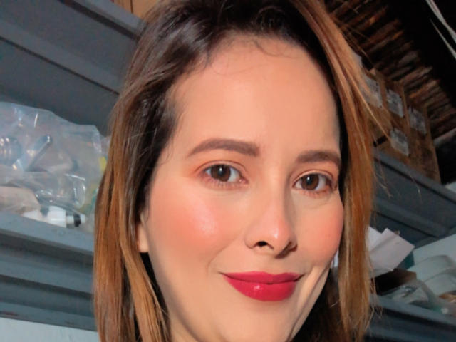 SaritaVelez has a cute face with brown eyes, pinkish skin, red lipstick, and brown hair, smiling slightly in a selfie. Her tits aren't visible.
