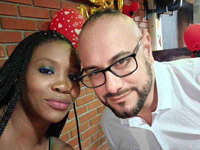 MaxAndMegan69 (dark-skinned woman with braids, blue eyeliner, pink lipstick, small boobs) and bald man with glasses, white shirt, and beard, smiling in selfie.