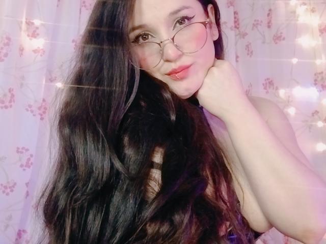 MillaDrew with long black hair, glasses, pink blush, and a cute smile, resting her chin on her hand, showing her bare shoulder and upper chest. Her tits are slightly visible.