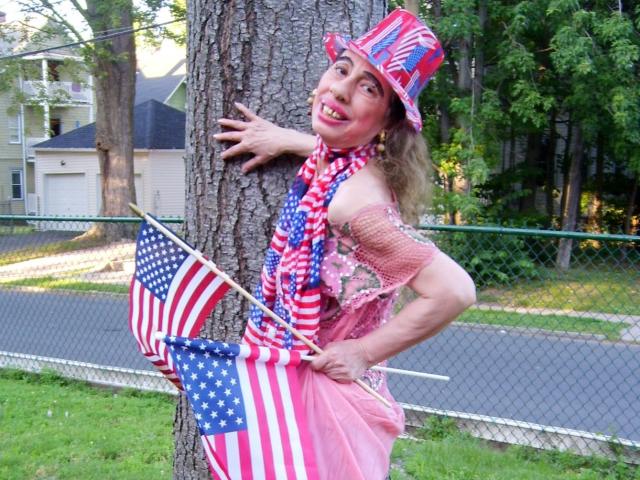 ElenahHottie draagt een rode, witte en blauwe hoed, een roze jurk en sjaal, en houdt twee Amerikaanse vlaggen vast. Ze staat tegen een boom, met een gekke glimlach en haar hand op de stam..