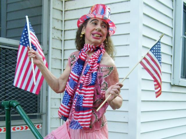 ElenahHottie porte un chapeau rouge, blanc et bleu, une écharpe assortie et une robe rose, tout en tenant des drapeaux américains dans chaque main, et se tient devant une maison blanche, souriant joyeusement..
