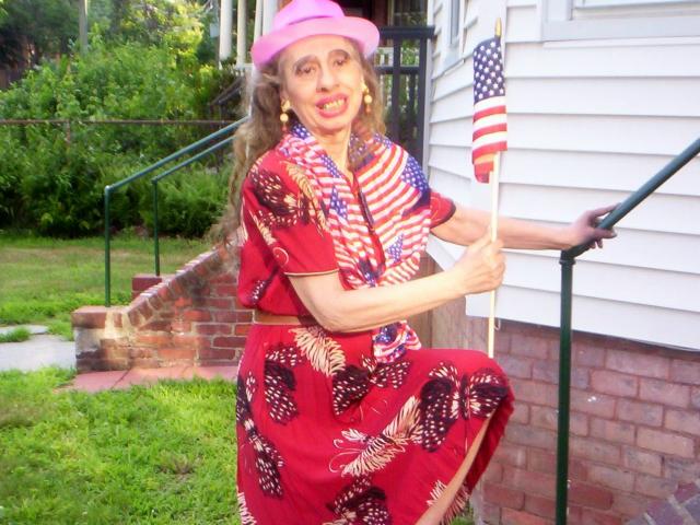 ElenahHottie in a red floral dress with American flag scarf, pink hat, holding tiny American flag, standing by white brick house, grass, and brick stairs, smiling, with yellow earrings.