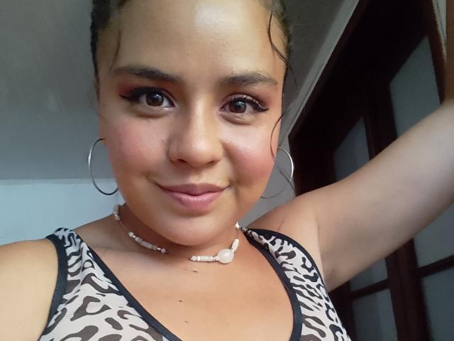 ArielleWilde with tan skin, brown eyes, and curly hair, wearing a leopard print top, hoop earrings, and a bead necklace, smiling at the camera. Her tits are small.