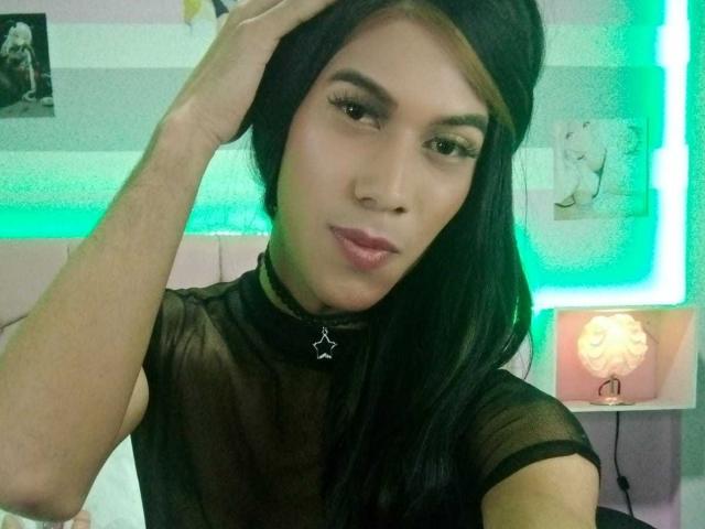 MitchellTay with long black hair, wearing a sheer black top, black choker, and pink lipstick, hand in hair, green neon light, and a cute face, looking at camera.