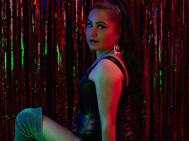 SussyFantasy with long black hair in a ponytail, wearing a black leather corset, knee-high silver glitter socks, and large hoop earrings, looking over shoulder in colorful light.
