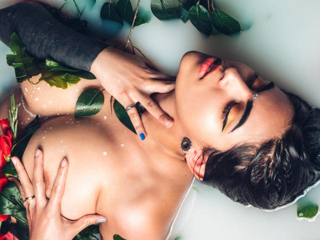 CoriPaige, com os olhos fechados, pele molhada e folhas verdes cobrindo os seios, segurando os seios, usando um piercing no nariz, em uma banheira com pétalas de rosa..