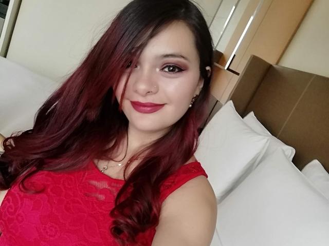 KatherinLatin with dark red hair, red lipstick, and a small nose piercing, smiling softly. She's got a cute face and brown eyes. Her tits aren't visible.