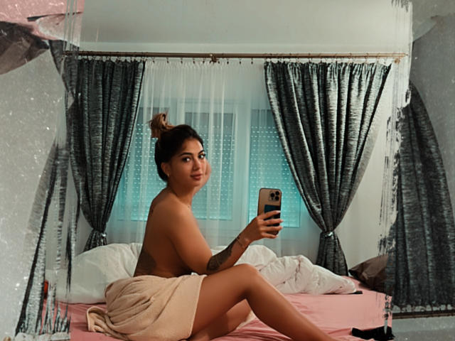 MorenaS, avec son corps bronzé et tatoué, les cheveux bruns attachés en chignon, et une serviette rose recouvrant ses fesses, assise sur un lit, prenant un selfie devant un miroir..