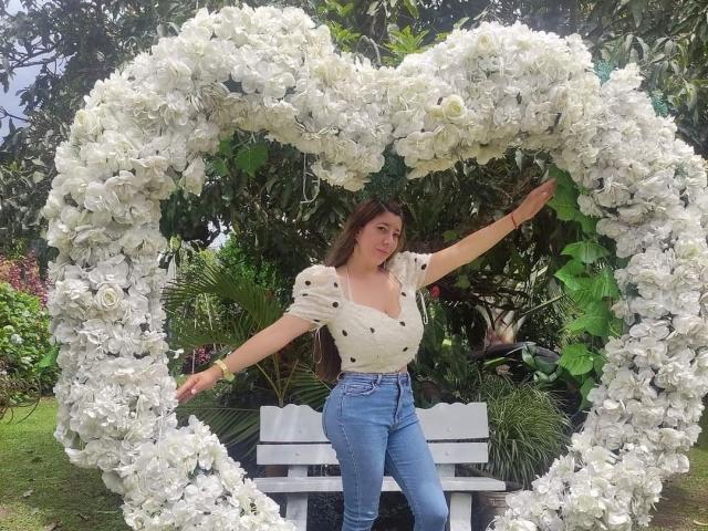 HotPamelaSex stands in front of a heart-shaped white flower arch, wearing a polka-dot top and blue jeans, with arms outstretched, in a lush garden.