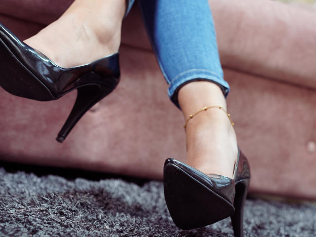 Les pieds de JulianaCastiblanco, chaussés de talons hauts noirs et brillants, avec des chevilles ornées d'une cheville dorée, portant un jean bleu, debout sur un tapis sombre et moelleux..