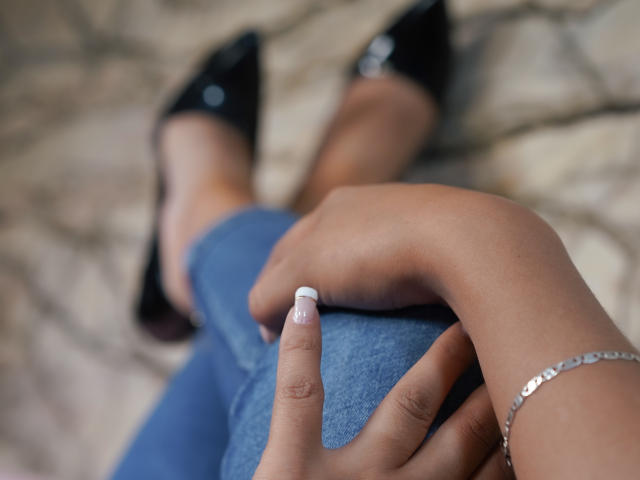 JulianaCastiblancos Hände mit weiß lackierten Nägeln ruhen auf ihrer blauen Jeans, sie trägt schwarze spitze High Heels, ein silbernes Armband an ihrem Handgelenk und hat eine braune Haut..