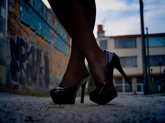JulianaCastiblancos glänzende schwarze High Heels mit dicken Stilettoabsätzen, stehend auf einem rauen Bürgersteig, Beine in dunklen Strumpfhosen, im Hintergrund eine städtische Graffiti-Wand, bewölkter Himmel..