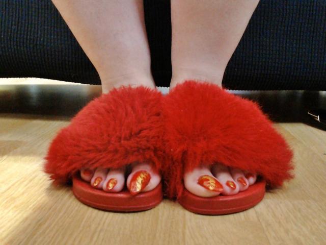 Os pés de KaryQueen estão usando pantufas vermelhas fofas, mostrando suas unhas dos pés pintadas de vermelho com glitter dourado, em um chão de madeira. Suas pernas nuas são visíveis acima das pantufas..