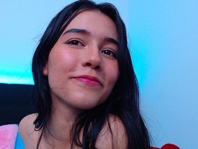 DeisyWest with long black hair, pink lips, and a slight smile, has a small zit on her right cheek, wearing a blue and pink top, in a blue-lit room.