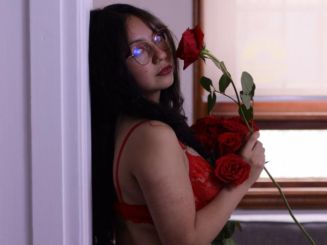 VeroHotLove with long black hair, red lace bra, and glasses, holding red roses, looking over shoulder with blue eye reflection, in dimly lit room. Boobs, roses, sexy.