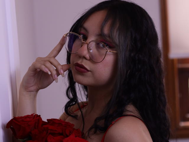 VeroHotLove, con cabello negro ondulado, lápiz labial rojo y gafas, tocándose la nariz, vestida con un vestido rojo con detalles de rosas, tiene un aspecto lindo y sexy..