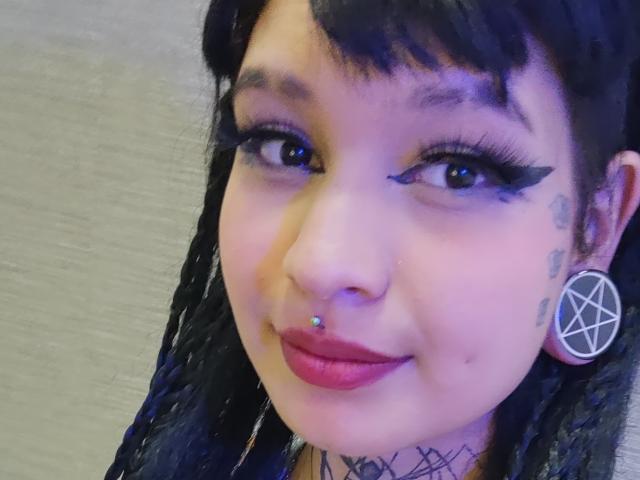 SayuriRose tiene trenzas oscuras, delineador de ojos negro, lápiz labial morado, un piercing en la nariz y un arete con un pentagrama, además de tatuajes en la mejilla y el cuello. Su sonrisa es linda..