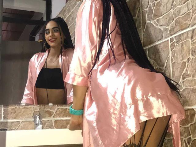 SexyHannaD with long black braids, pink satin robe, black crop top, and black fishnet stockings, leans on a sink, smiling at her reflection in a mirror.