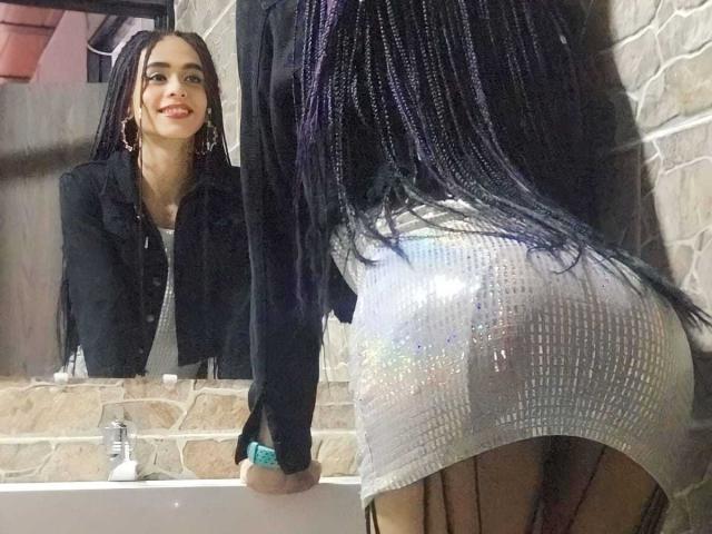 SexyHannaD with long purple braids, black jacket, and silver sparkly skirt, leaning on bathroom sink, looking in mirror, showing her shiny ass, smiling, wearing hoop earrings.