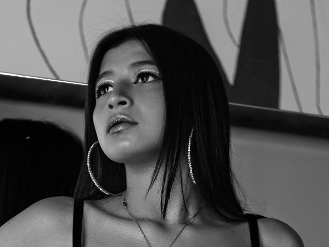NaiaSanders with dark, straight hair, wearing large hoop earrings and a thin necklace, has shiny lips and a slight smile, looking upward in a black-and-white photo.