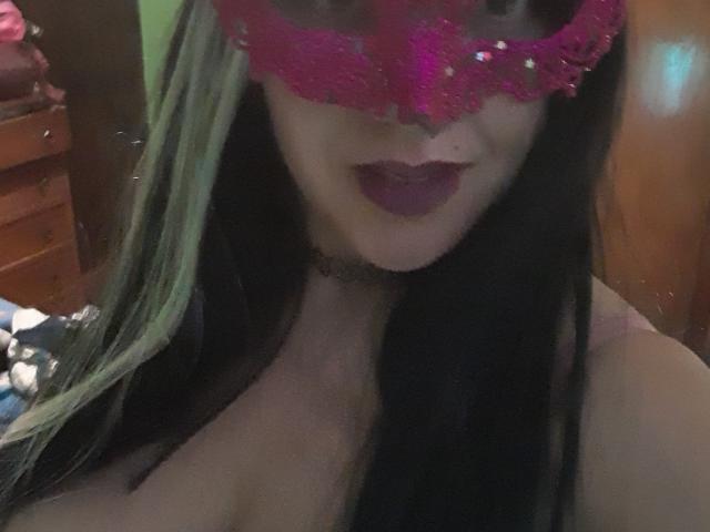 HotNata, con i suoi lunghi capelli neri, la mascherina di pizzo rosa, il rossetto viola scuro, la collana nera e il décolleté evidente, mostra il suo viso e il suo seno sensuali in una stanza poco illuminata..