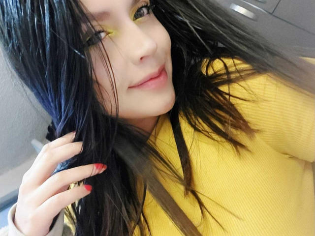LinaStars with wet black hair, yellow eyes, and red nails, wearing a yellow top, smiling slightly. Her hand touches her hair. She has pale skin and small tits.