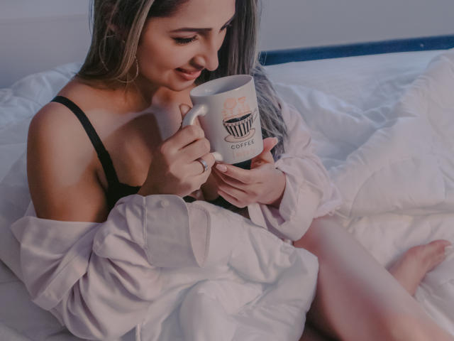 StefanyCaruso with tits visible in a black bra, holding a white coffee mug, sitting on a bed with white sheets, wearing an off-shoulder pink shirt.