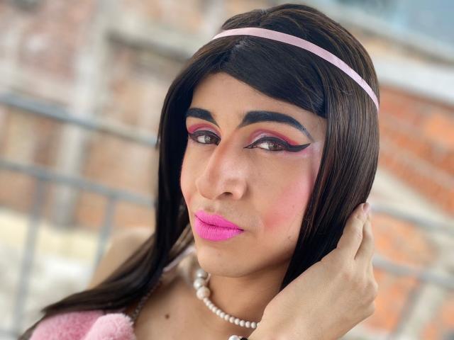 Catalina Castro tiene lápiz labial rosa, delineador de ojos llamativo y una diadema rosa. Está tocándose la cabeza con un collar de perlas. Sus senos son pequeños y su trasero no se ve..