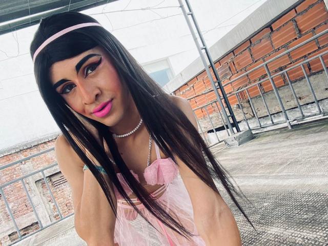 Catalina Castro heeft lang donker haar, roze eyeliner en roze lippenstift. Ze draagt een roze jurk, een hoofdband en een ketting, en leunt voorover met een schattende glimlach. Haar borsten zijn zichtbaar..